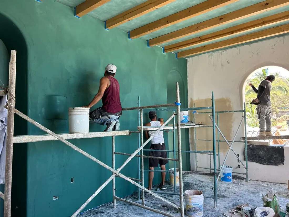 Dos aplicadores de ArquiVal extendiendo pasta en color verde azulado sobre un muro interior de doble altura, subidos en andamio