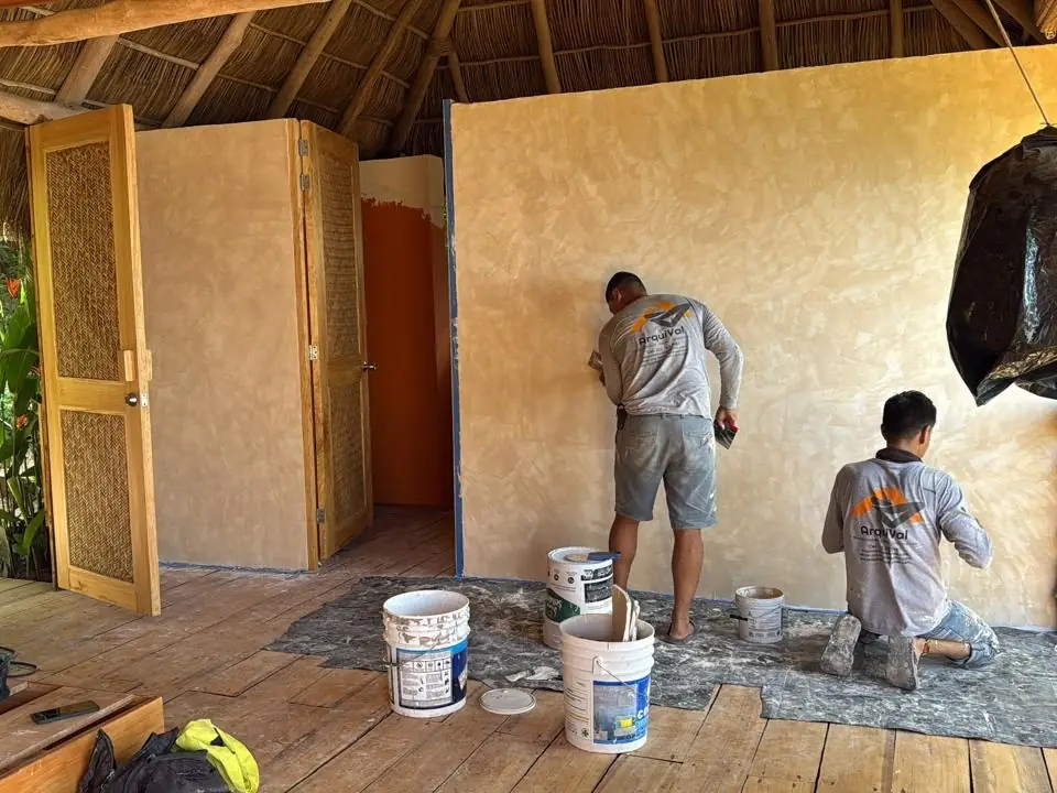 Dos aplicadores de ArquiVal extendiendo pasta Corev en tono ocre sobre un muro, bajo una techumbre de palapa con puertas de madera