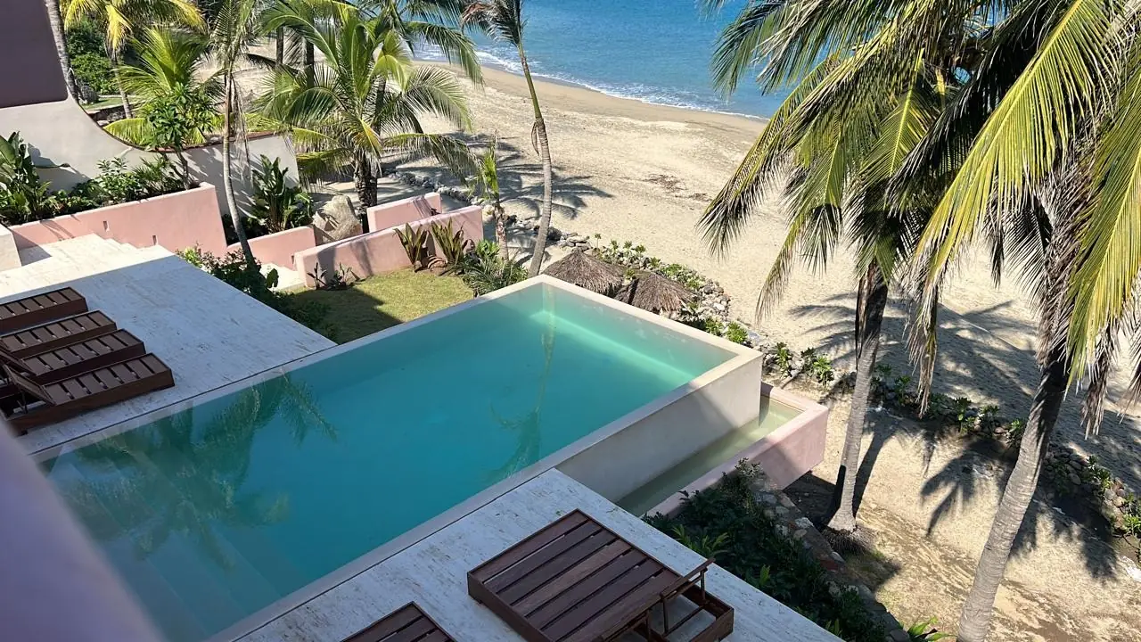 Infinity pool finished in chukum overlooking the ocean, the water reading green-turquoise over the material’s pale ochre base, with palms and beach beyond